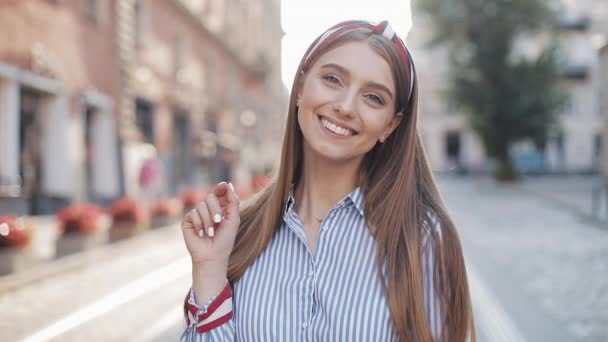 Portrait of Young Happy Attractive Girl with Straight Brown Hair and Blue Eyes Wearing in Striped Dress and Fashionable Headband Smiling Looking to Camera Waving Hi Standing at the City Street Zoom. - Felvétel, videó