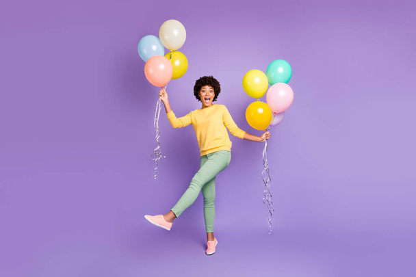 Full size photo of funky funny teen mulatto girl hols many colorful baloons feel content crazy on spring holidays wear green sweater green pants trousers isolated over violet purple color background - Photo, Image