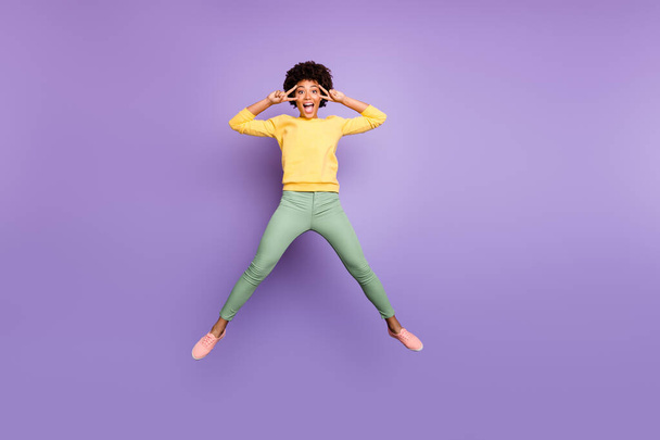Full body photo of amazed carefree afro american girl jump have fun on autumn spring holidays make v-signs wear yellow pullover green pants trousers isolated over violet purple color background - Foto, immagini