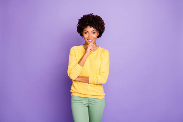 Photo of cheerful puzzled cute girl wearing yellow clothing posing in front of camera touching her chin smiling toothily beaming isolated over purple pastel color background - Foto, imagen