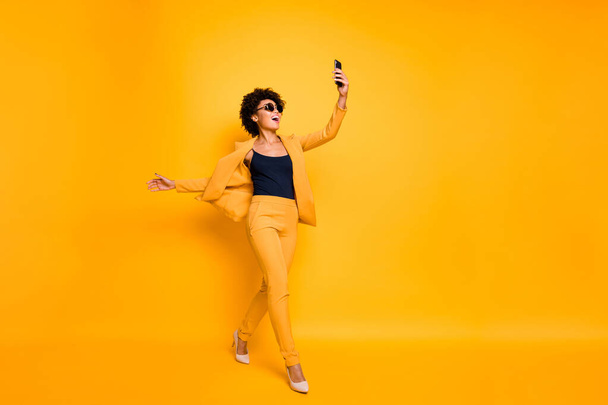 Full body profile side photo of cheerful wavy hair girl have fun on leisure time go take selfie feel content wear style trousers sunglass blazer high-heels isolated yellow color background - Photo, Image