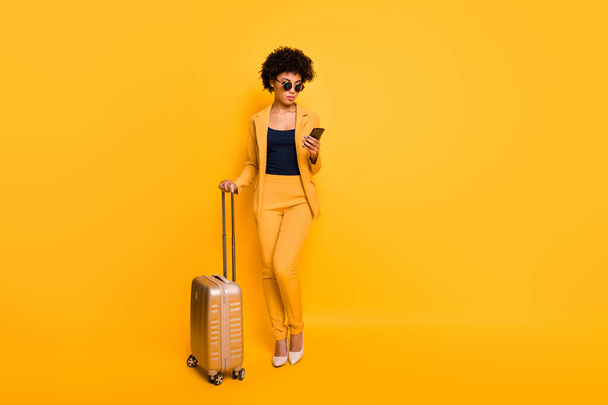 Full length photo of focused brown curly hair girl use cellphone search taxi hold bags tourism concept have abroad vacation wear style jacket pants high-heels isolated over yellow color background - Photo, Image