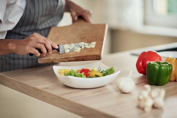 Cerrar mujer de raza mixta en delantal sosteniendo tabla de cortar y poner el ajo picado en un tazón con verduras
. - Foto, imagen