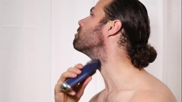 Uzun saçlı yakışıklı genç adam banyoda tıraş makinesiyle sakalını tıraş ediyor. Güzel ışık, bulanık arkaplan. - Video, Çekim