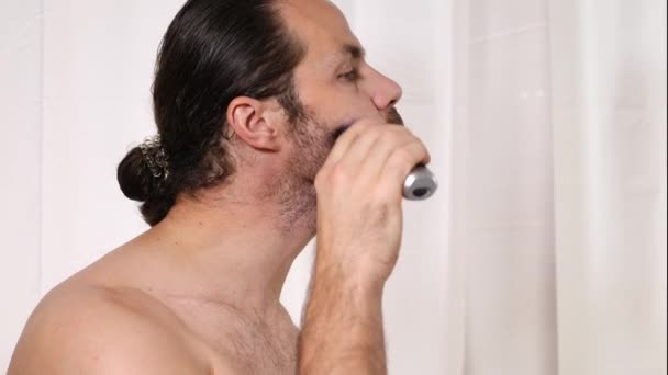Un joven guapo con el pelo largo se afeita la barba con una podadora en el baño. Hermosa luz, fondo borroso
. - Imágenes, Vídeo