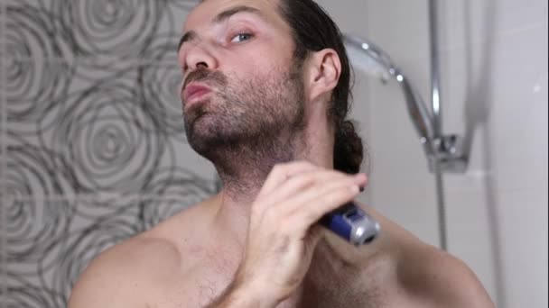 Uzun saçlı yakışıklı genç adam banyoda tıraş makinesiyle sakalını tıraş ediyor. Güzel ışık, bulanık arkaplan. - Video, Çekim