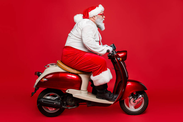 Profile side view portrait of nice bearded serious confident Santa father riding motor bike delivering fairy tale dream shopping isolated on bright vivid shine vibrant red color background - Foto, Imagem