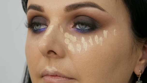 Face contouring makeup artist leads a woman in the face with a brush for applying powder, foundation or concealer. Lilac and pearly smoky eyes eyeshadow, eyes and face of a woman close up - Footage, Video