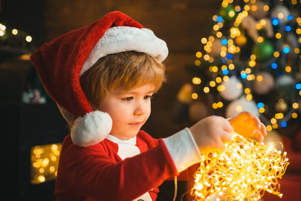 Child with garland lights at Christmas tree and fireplace on Xmas eve. It is miracle. Family with kids celebrating Christmas at home. Boy cute child cheerful mood play near christmas tree. - Zdjęcie, obraz