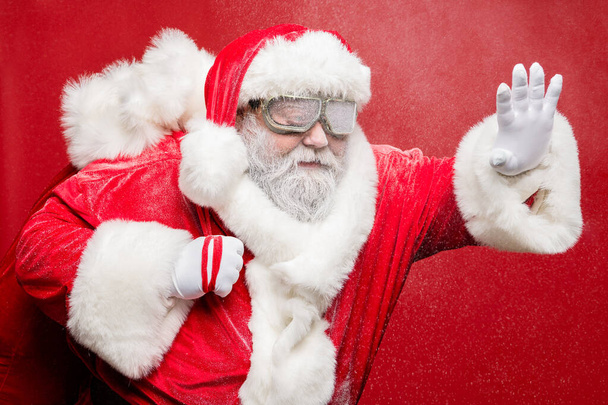 Профиль боковой фото концентрированного престарелых Санта Клаус держать его за руку имеют дорожные проблемы в катаклизме снежной бури нести сумку мешок носить защитные очки колпачок шляпу перчатки изолированы на красном фоне
 - Фото, изображение