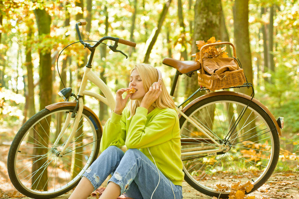Dreamy young college girl relaxing in the park eating croissant and listening thoughtfully to music in headphones. Active leisure and lifestyle. Girl ride bicycle for fun. - Photo, Image