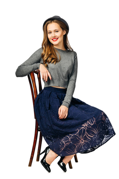 young beautiful girl in dark blue skirt and gray jumper, and gray hat sitting on old chair. isolated on white background - Foto, Imagen