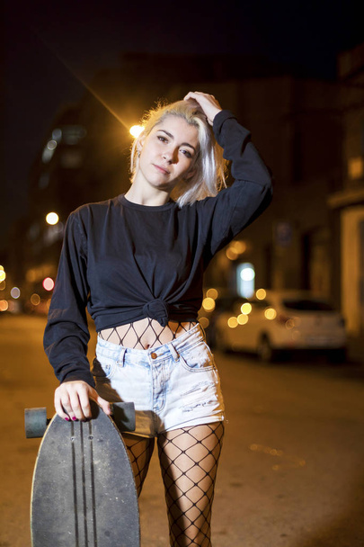Vooraanzicht van jonge mooie vrouw op zoek camera in korte broek terwijl staande op de straat houden een skateboard 's nachts in de stad - Foto, afbeelding