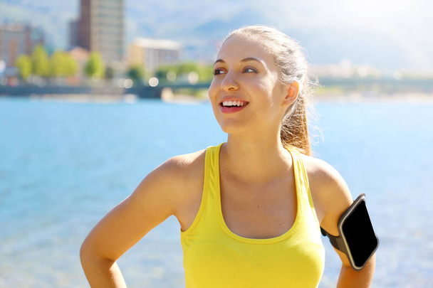 Счастливая фитнес-женщина в спортивной одежде и спортивной повязке для смартфона пустой для рекламы бега или кардио тренировки на пляже
. - Фото, изображение