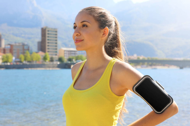 Successful positive female athlete wearing armband blank for advertising before running or exercising outdoor in summer. Woman success in sport lifestyle. - Фото, зображення