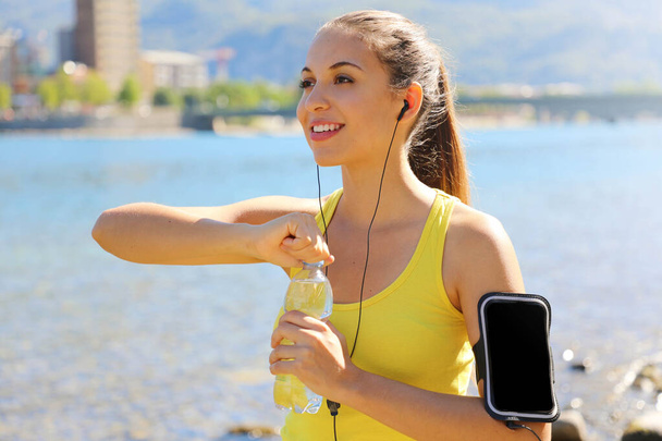 Thirsty fitness woman opens bottle of water after training outdoor. Fit woman using smartphone fitness app on armband as activity tracker. - Φωτογραφία, εικόνα