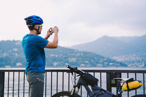 Cyclist taking pictures with smart phone. Caucasian man in bike helmet taking photo of landscape. Outdoor activity. Tourist makes photo of Lake Como in Italy - Photo, Image