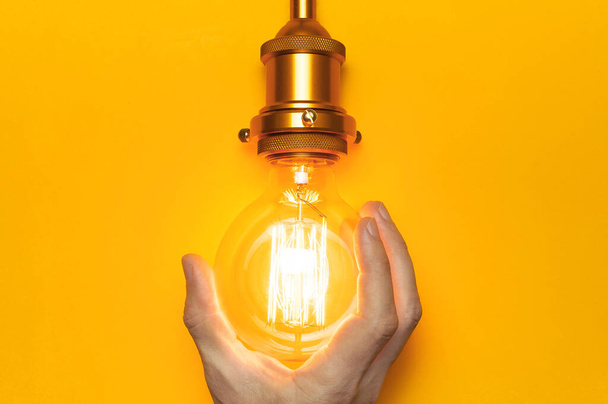 Винтажный модный Edison lamp в мужских руках на ярко-желтом фоне. Плоское копировальное пространство с видом сверху. Концепция креативной идеи, дизайнерский ламп, современный предмет интерьера. Освещение, электричество
 - Фото, изображение
