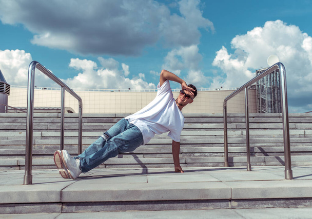dancer on one arm dances break dance, hip-hop. In summer, city background steps clouds. Active youth lifestyle, young male dancer, fitness movement workout breakdancer jeans sneakers, sunglasses. - Photo, Image