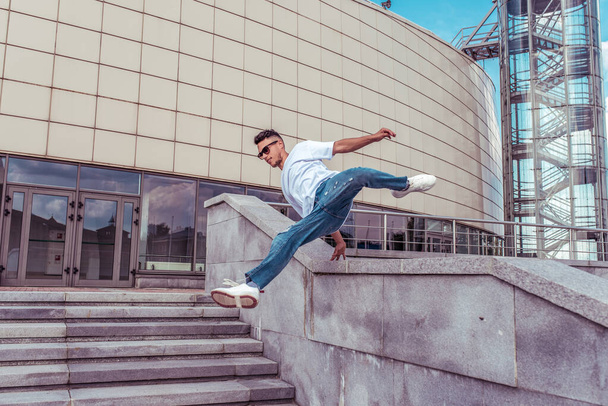 Dancer jumping jump dancing break dance, hip hop. Summer city, background building. Active youth lifestyle, young male dancer, fitness movement workout breakdancer. In jeans and sneakers, sunglasses. - Photo, image