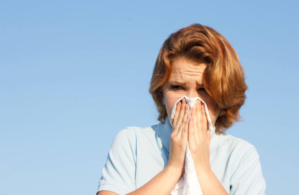 Young woman blowing her nose into handkerchief or tissue at summer sunny hot day. Sick girl suffering from runny nose, snot, allergy, allergic reaction. Health, medical concept on blue sky background. - Foto, imagen