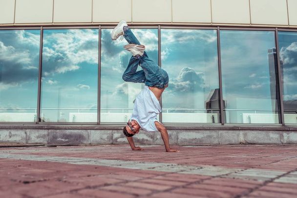Handstand dancer dancing break dance, hip hop. In summer city, background is glass windows. Active youth lifestyle, young male dancer, fitness workout. In jeans sneakers, sunglasses. Free space text. - Zdjęcie, obraz