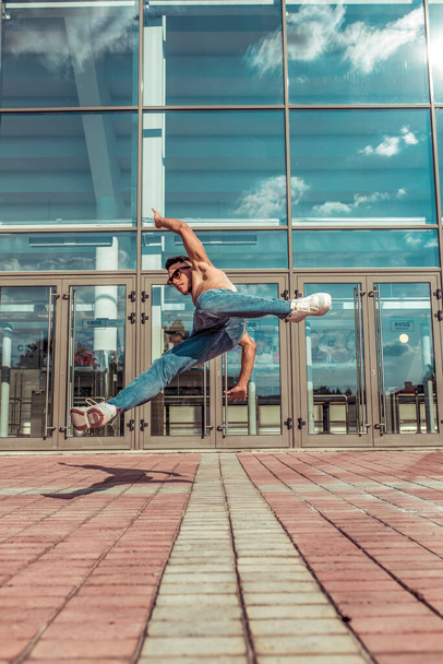 Jumping dancer, acrobat actor break dance, hip hop. In summer city, background is glass windows clouds. Active youth lifestyle, young male dancer, fitness movement workout breakdancer. - Fotó, kép