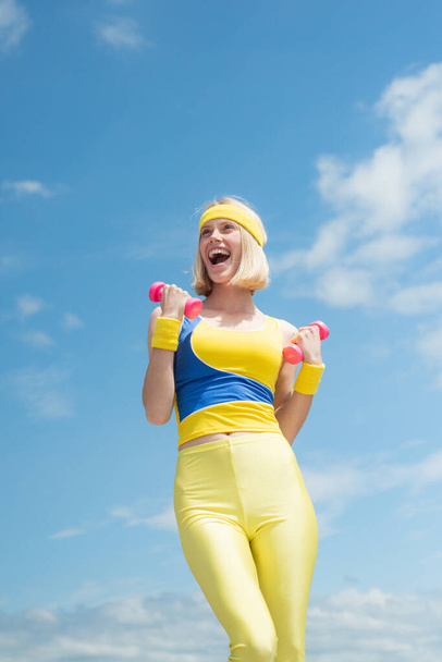 Successful attractive sportswoman with bright smile wearing yellow with blue training uniform demonstrating exercises with pink dumbbells on sky background with clouds. Fashion and Trends in Sport - Photo, Image