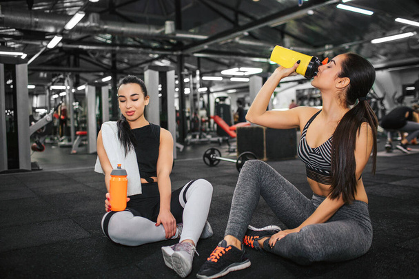 Calm and peaceful young women sit on floor in gym. Asian young model look down at orange water bottle. Her friend drinks from yellow one. They alone in gym. - Fotoğraf, Görsel