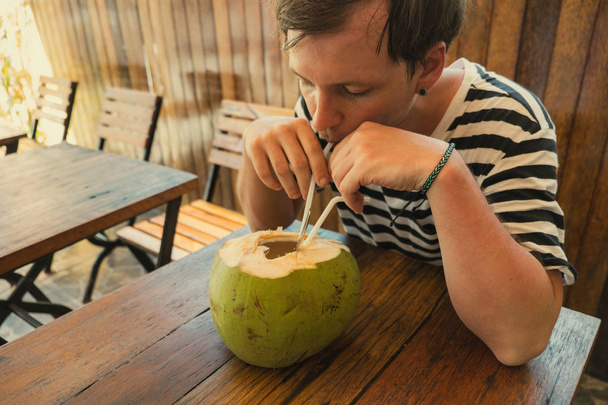 Młody człowiek pije świeże kokosy przy stole w kawiarni. Trav żywnościowy - Zdjęcie, obraz