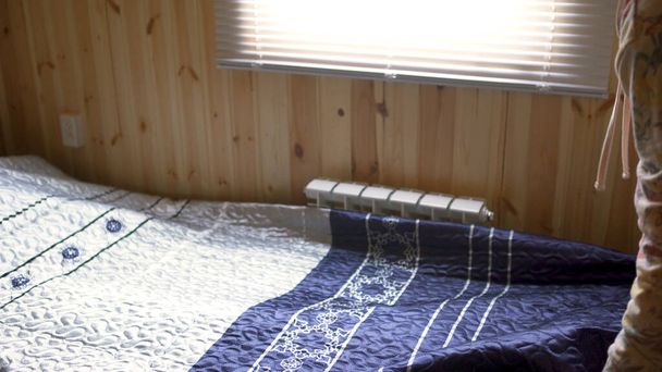 Вид сбоку женщины, делающей кровать в маленькой комнате с деревянными стенами и ярким солнцем из окна. Искусство. Женщина расстелила одеяло по узкой кровати внутри деревянного дома
. - Фото, изображение