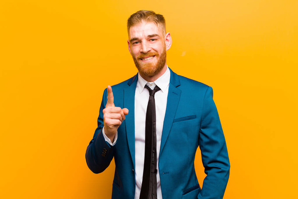 young red head businessman smiling and looking friendly, showing number one or first with hand forward, counting down against orange background - Foto, Imagen