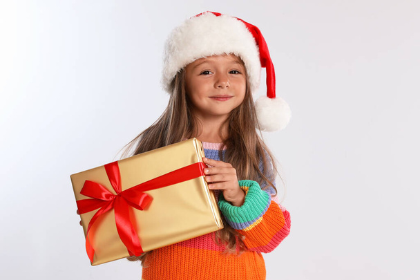 Happy little child in Santa hat with gift box on light grey background. Christmas celebration - Photo, Image