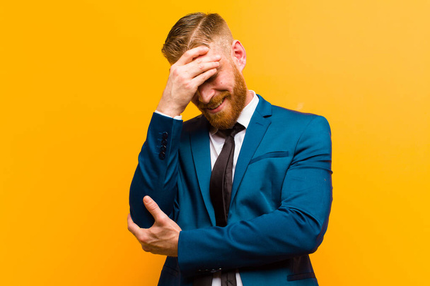 jonge rode hoofd zakenman kijken gestrest, beschaamd of boos, met een hoofdpijn, het bedekken van gezicht met de hand tegen oranje achtergrond - Foto, afbeelding