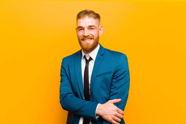 young red head businessman laughing shyly and cheerfully, with a friendly and positive but insecure attitude against orange background - Fotografie, Obrázek