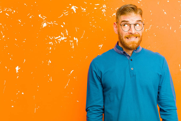 young red head man looking worried, stressed, anxious and scared, panicking and clenching teeth against grunge orange wall - Photo, Image
