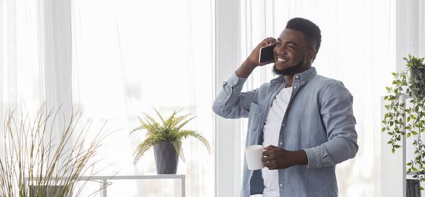 Усміхнений хлопець розмовляє на мобільному телефоні і п'є каву біля вікна
 - Фото, зображення