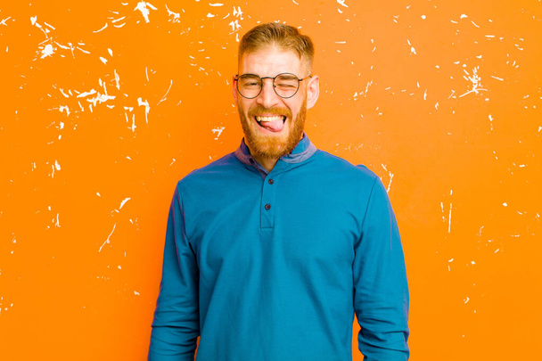 young red head man with cheerful, carefree, rebellious attitude, joking and sticking tongue out, having fun against grunge orange wall - Zdjęcie, obraz