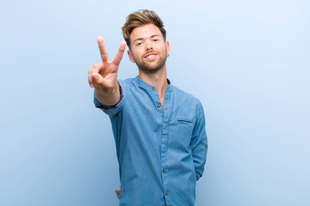 young businessman smiling and looking happy, carefree and positive, gesturing victory or peace with one hand against blue background - Фото, зображення