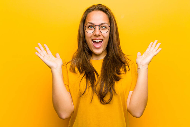 young pretty woman looking happy and excited, shocked with an unexpected surprise with both hands open next to face against orange background - Fotografie, Obrázek