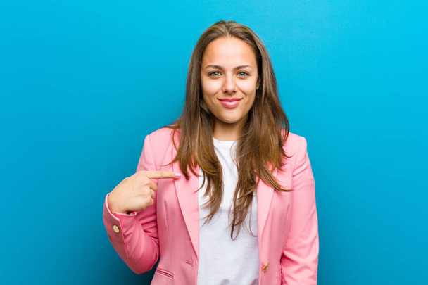 young woman looking proud, confident and happy, smiling and pointing to self or making number one sign against blue background - Fotografie, Obrázek