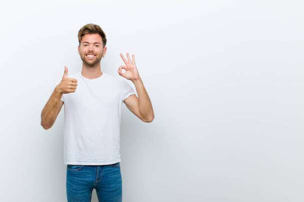 young man feeling happy, amazed, satisfied and surprised, showing okay and thumbs up gestures, smiling against white background - Photo, image