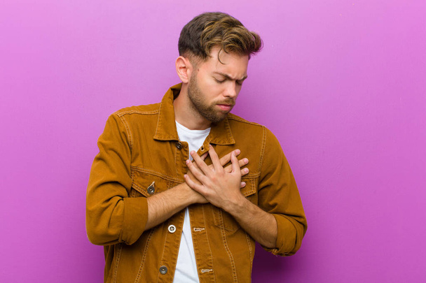 young man looking sad, hurt and heartbroken, holding both hands close to heart, crying and feeling depressed against purple background - Photo, Image