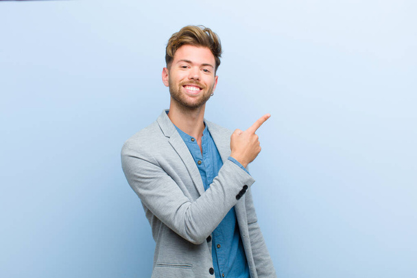 young businessman looking excited and surprised pointing to the side and upwards to copy space against blue background - Fotografie, Obrázek