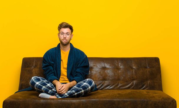 young man wearing pajamas looking puzzled and confused, biting lip with a nervous gesture, not knowing the answer to the problem . sitting on a sofa - Photo, Image