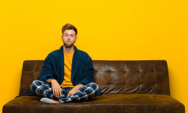 young man wearing pajamas feeling clueless, confused and uncertain about which option to pick, trying to solve the problem . sitting on a sofa - Photo, Image