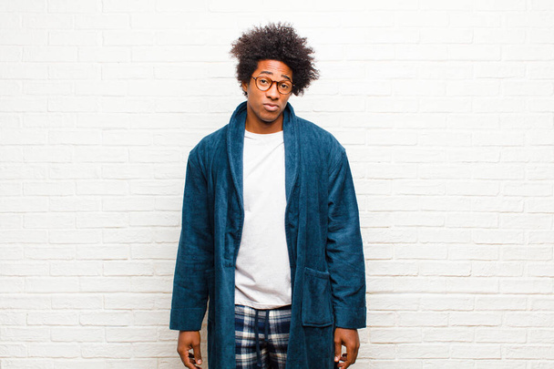 young black man wearing pajamas with gown feeling sad and whiney with an unhappy look, crying with a negative and frustrated attitude against brick wall - Photo, Image