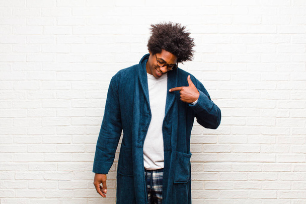 young black man wearing pajamas with gown smiling cheerfully and casually, looking downwards and pointing to chest against brick wall - Foto, Bild