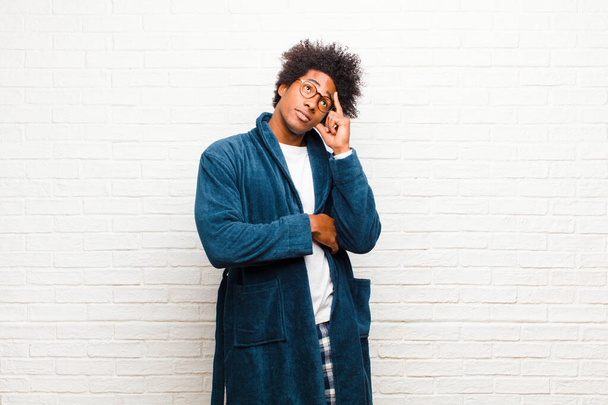 young black man wearing pajamas with gown with a concentrated look, wondering with a doubtful expression, looking up and to the side against brick wall - Foto, Imagem