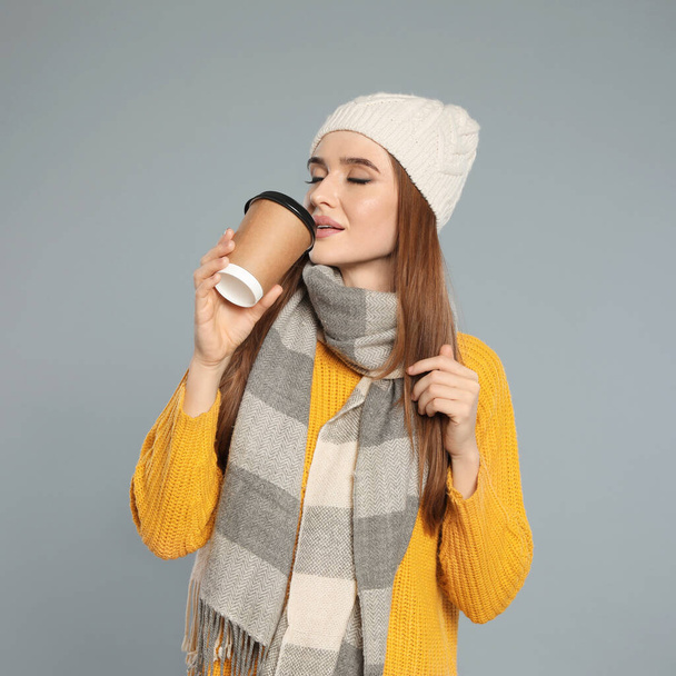 Gelukkige jonge vrouw in warme kleren met drank op grijze achtergrond. Winterseizoen - Foto, afbeelding